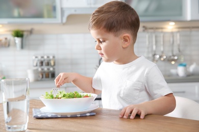 Photo of Adorable little boy eating vegetable salad at table in kitchen