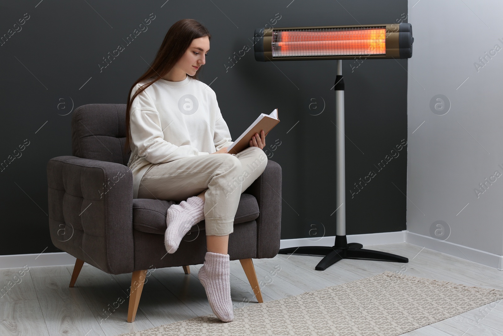 Photo of Young woman reading book on armchair near electric infrared heater at home. Space for text