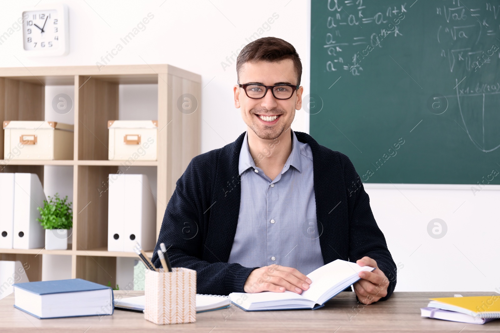 Photo of Young male teacher with book sitting at table in classroom