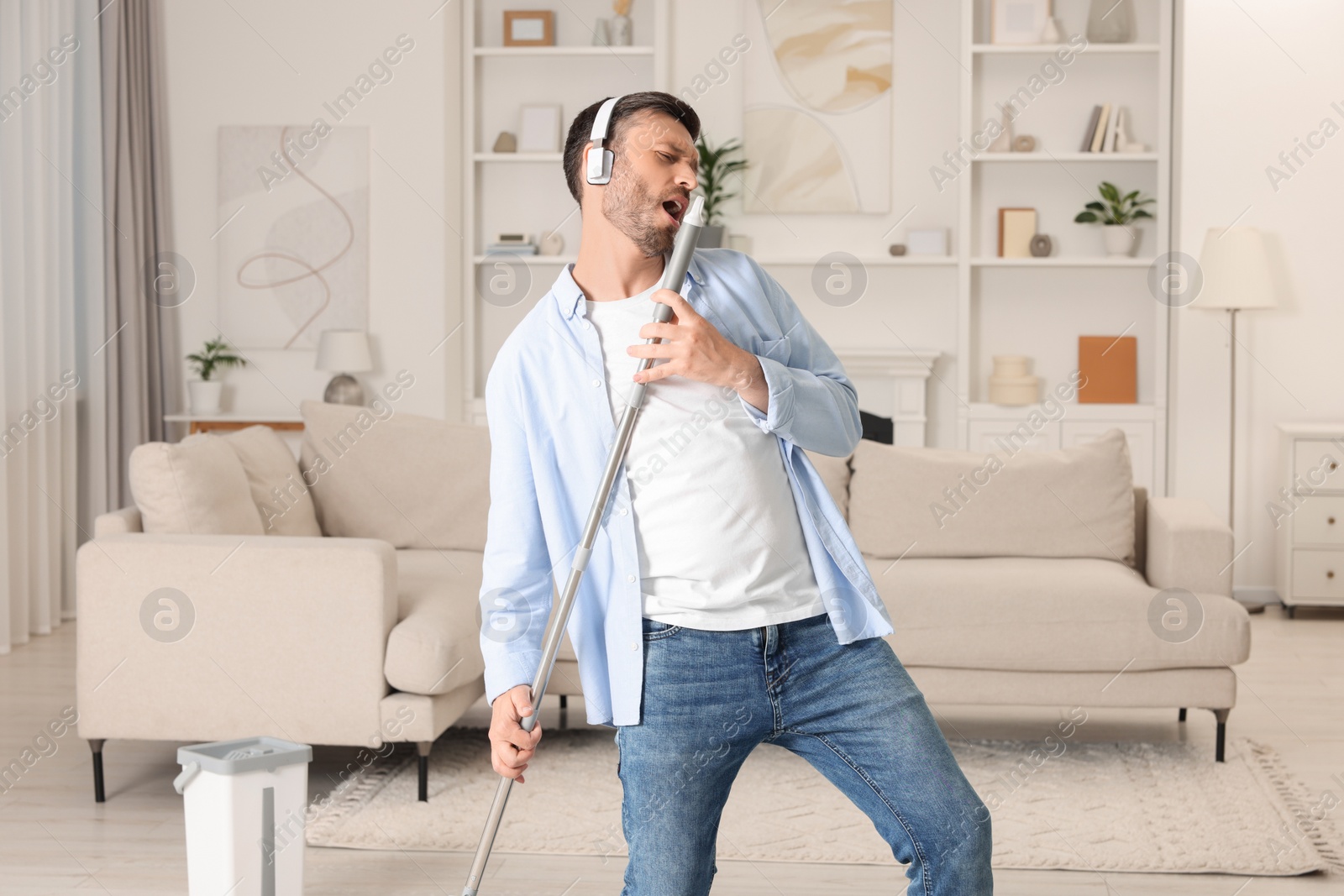 Photo of Enjoying cleaning. Happy man in headphones with mop singing while tidying up at home