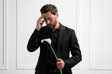 Sad man with calla lily flower mourning near white wall. Funeral ceremony
