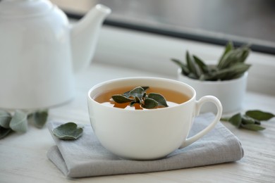 Photo of Cup of sage tea and green leaves on white wooden windowsill