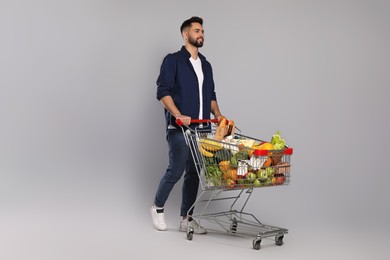 Photo of Happy man with shopping cart full of groceries on light grey background