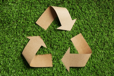 Photo of Recycling symbol cut out of kraft paper on green grass, top view