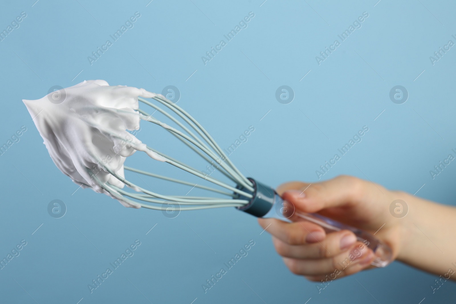 Photo of Woman holding whisk with whipped cream on light blue background, closeup