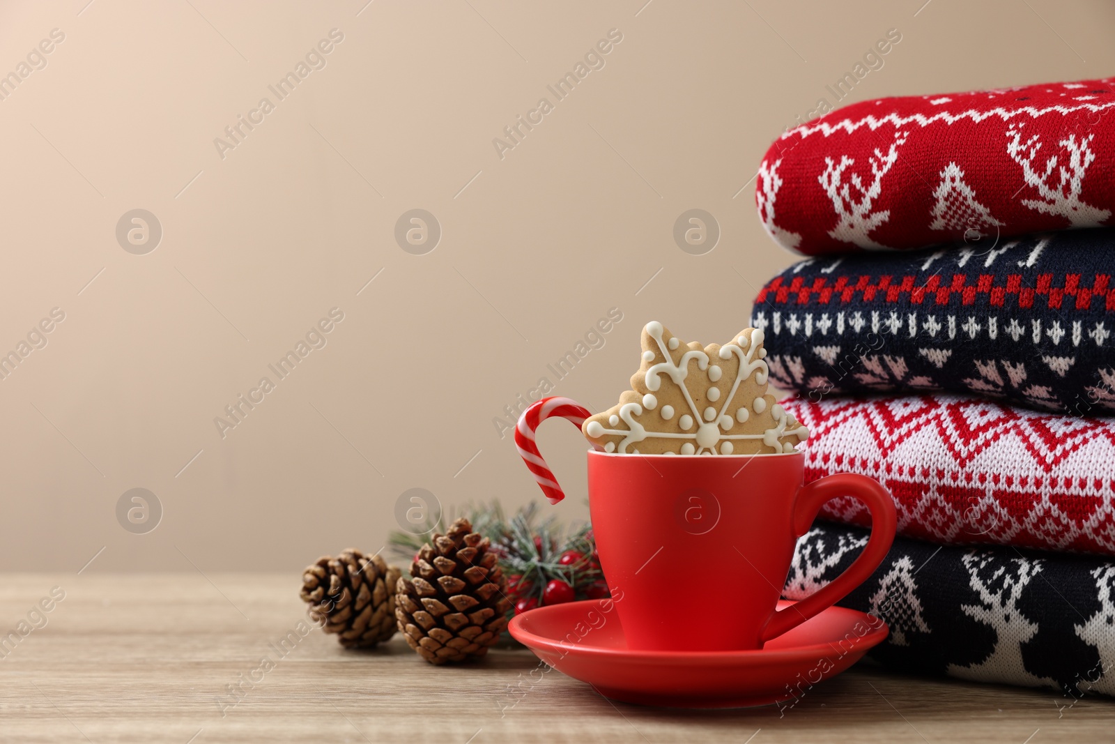 Photo of Stack of different Christmas sweaters, cup with candy cane, cookie and festive decor on wooden table, space for text