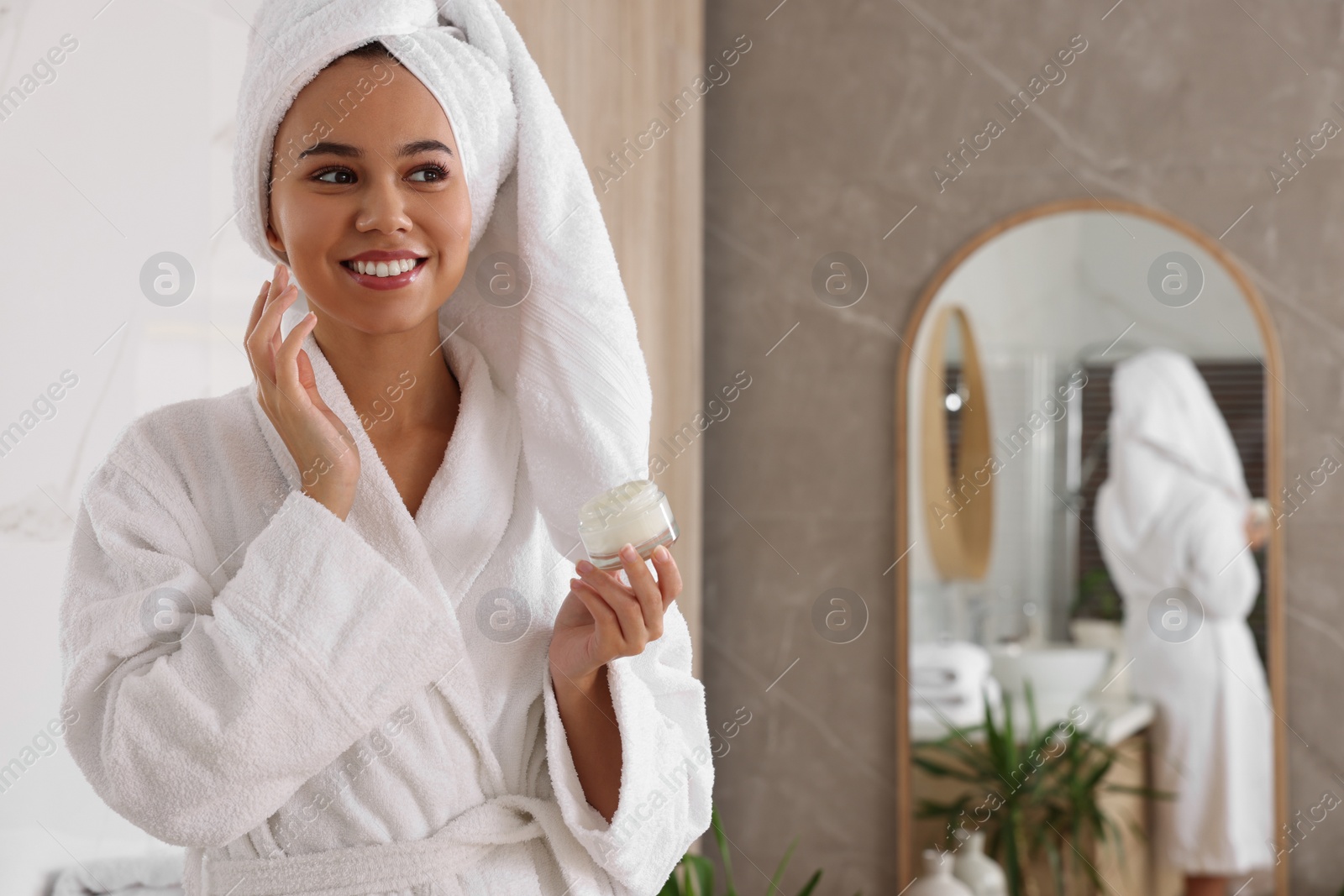 Photo of Beautiful African American woman applying cream onto face in bathroom, space for text