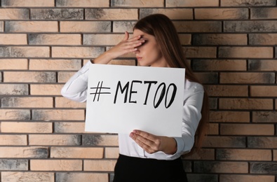 Woman holding paper with text "#METOO" near brick wall. Problem of sexual harassment at work