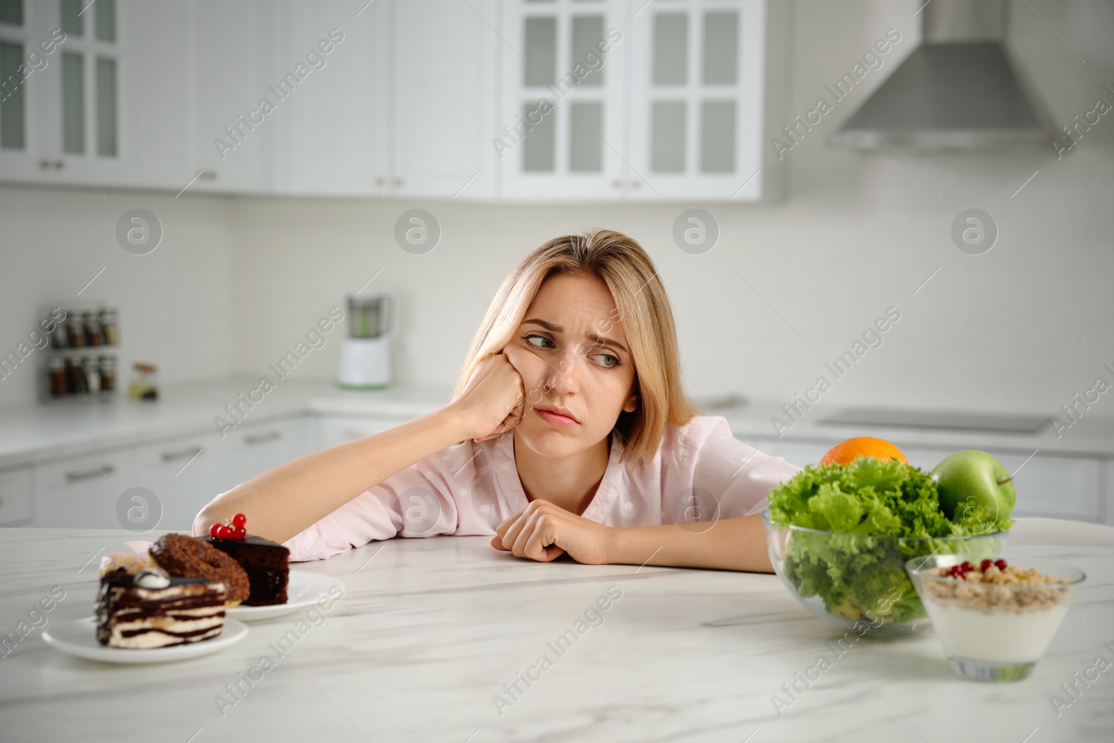 Photo of Woman choosing between sweets and healthy food at white table in kitchen