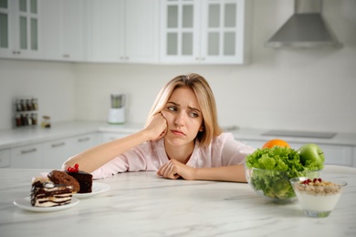 Woman choosing between sweets and healthy food at white table in kitchen