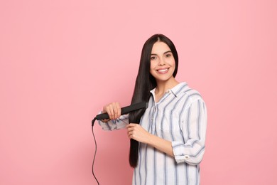 Beautiful happy woman using hair iron on pink background