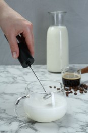 Woman whisking milk in cup with mini mixer (milk frother) at white marble table, closeup