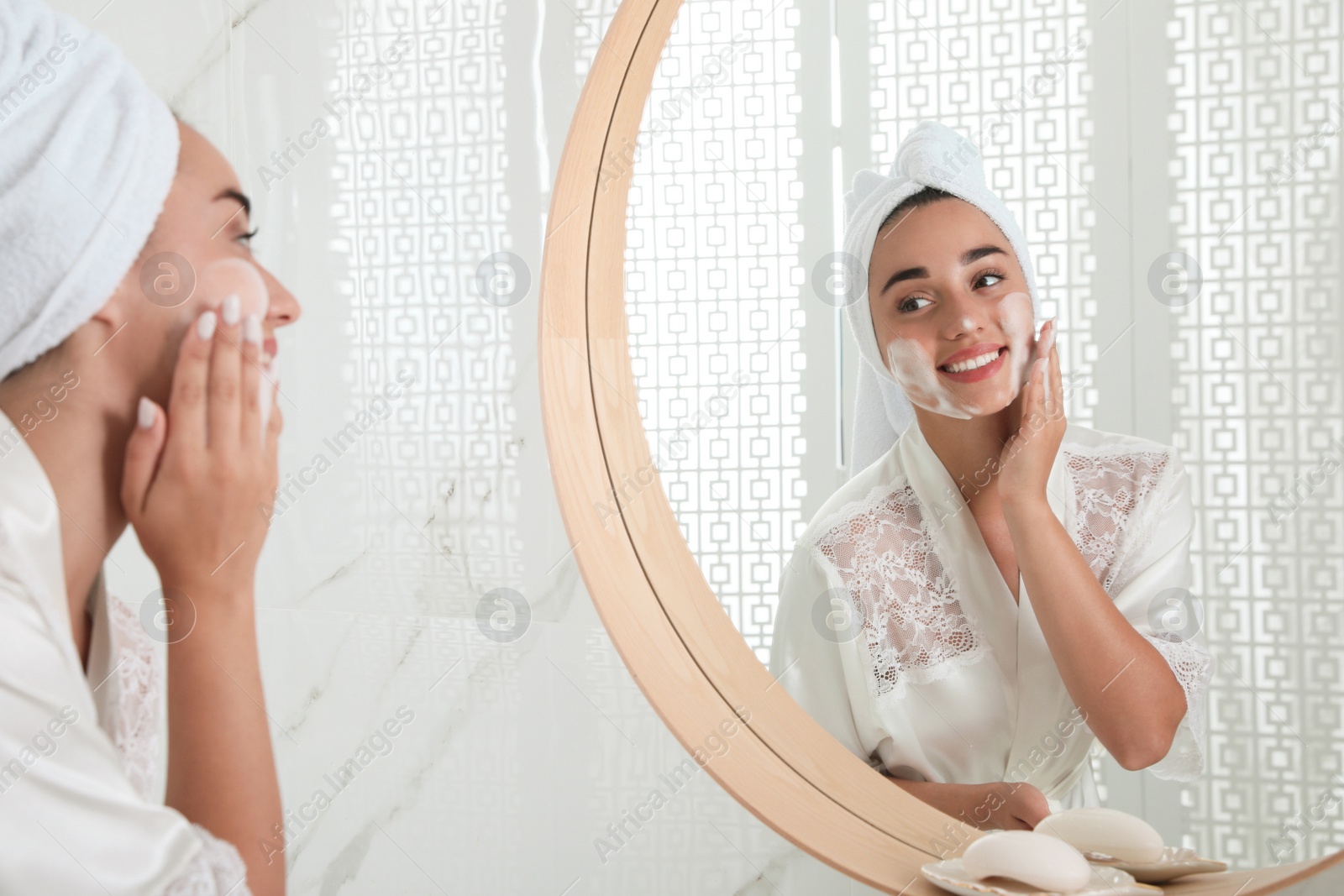 Photo of Beautiful young woman applying cleansing foam onto face near mirror in bathroom. Skin care cosmetic