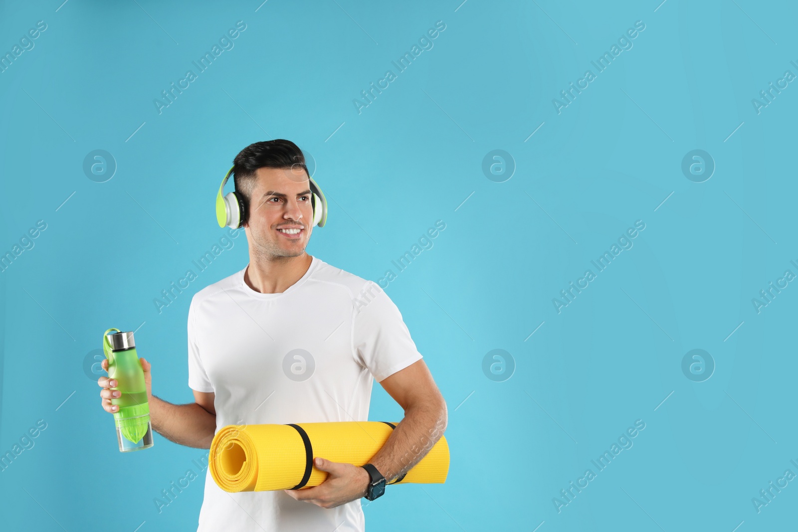 Photo of Handsome man with yoga mat, headphones and bottle on turquoise background. Space for text