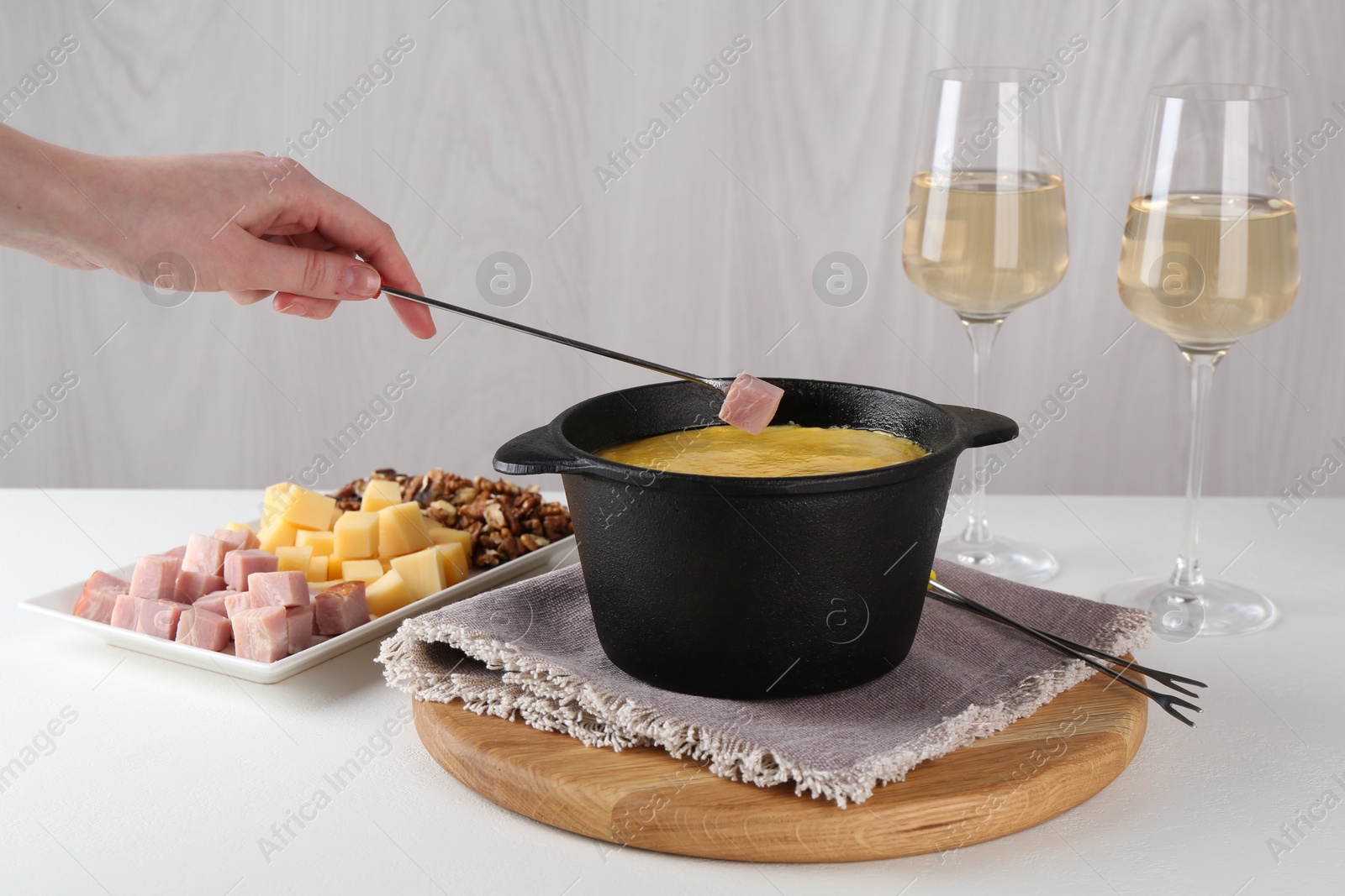 Photo of Dipping piece of ham into fondue pot with tasty melted cheese at white table, closeup