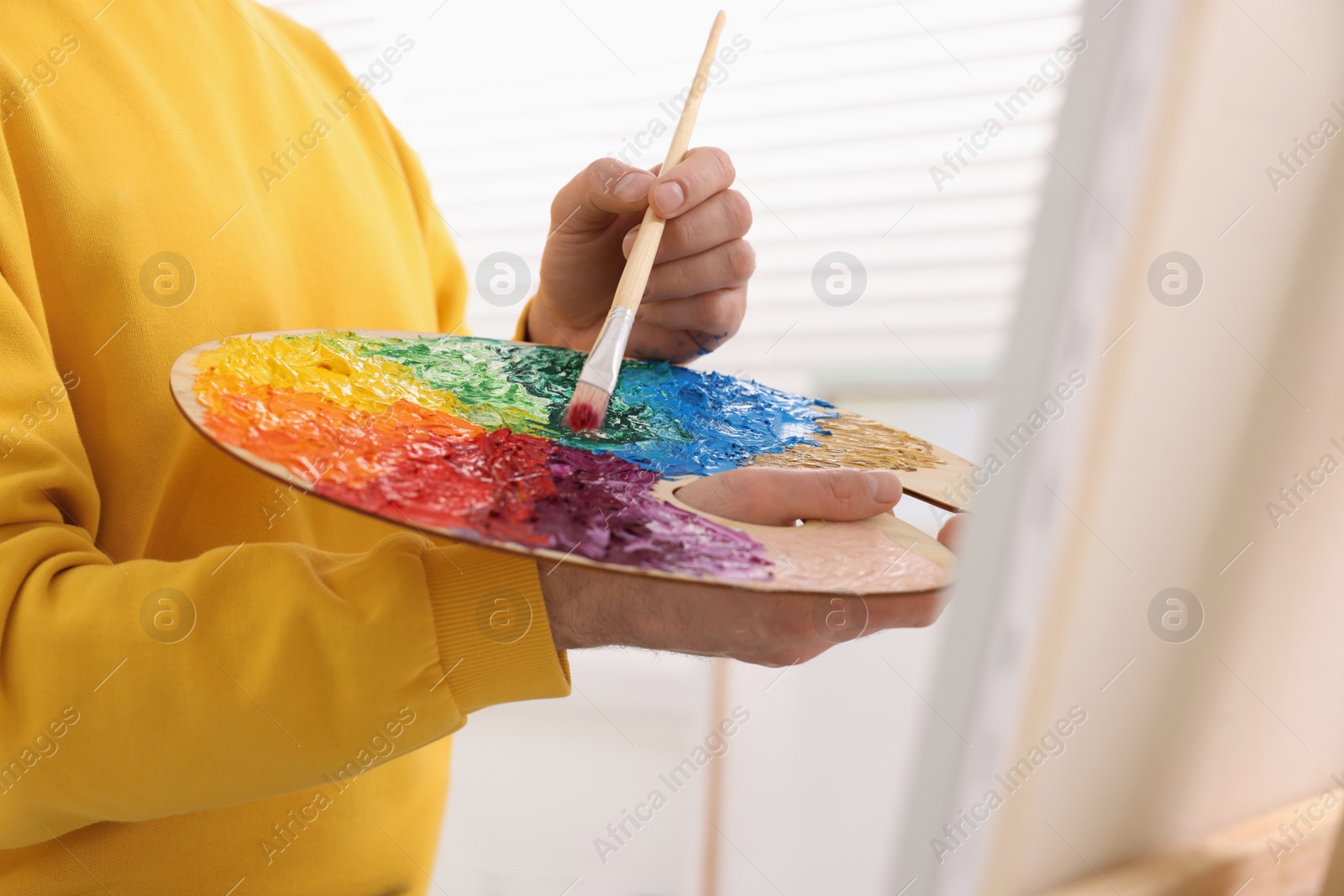 Photo of Man mixing paints on palette with brush near canvas in studio, closeup