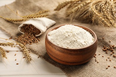 Photo of Wooden bowl of flour, wheat ears and sackcloth on white table