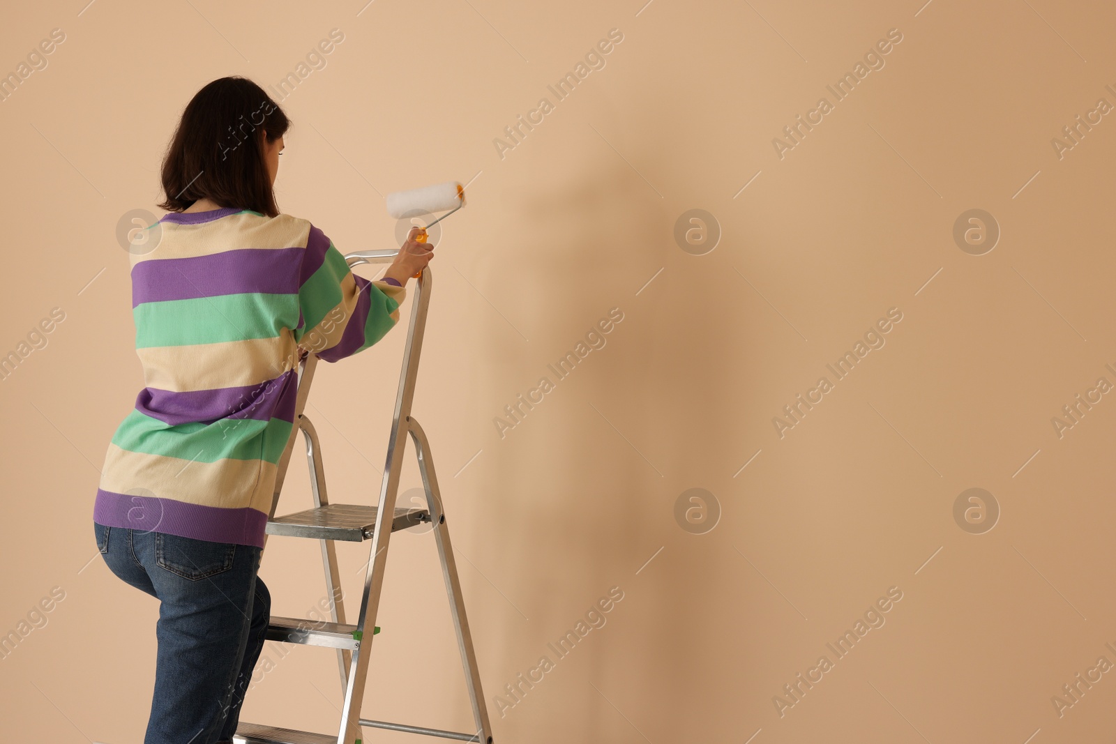 Photo of Young woman with roller on stepladder near pale orange wall indoors. Space for text