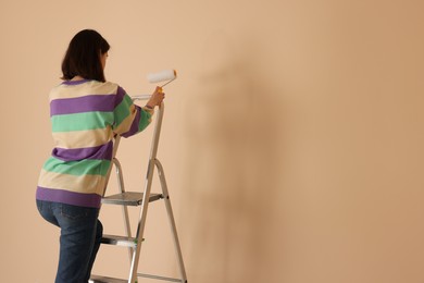 Photo of Young woman with roller on stepladder near pale orange wall indoors. Space for text