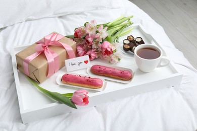 Photo of Tasty breakfast served in bed. Delicious desserts, tea, flowers, gift box and card with phrase I Love You on tray