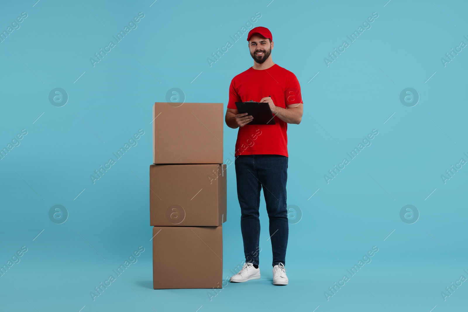 Photo of Happy courier with clipboard and stack of parcels on light blue background