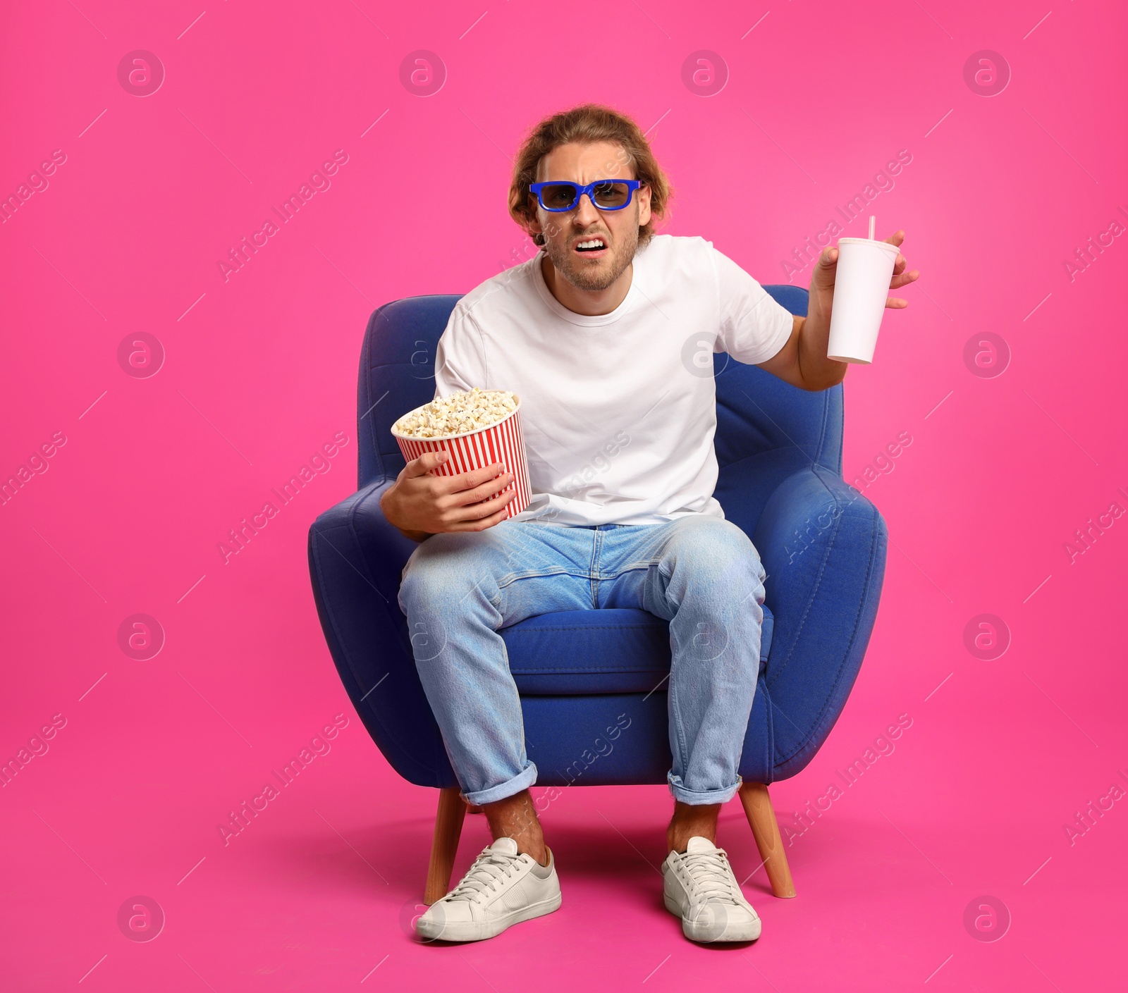 Photo of Emotional man with 3D glasses, popcorn and beverage sitting in armchair during cinema show on color background