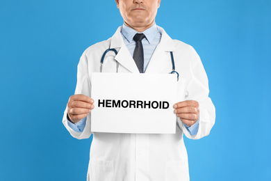 Doctor holding sign with word HEMORRHOID on blue background, closeup