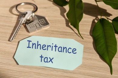 Photo of Inheritance Tax. Card, key with key chain in shape of house and green leaves on wooden table, closeup