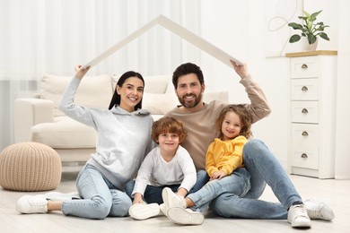 Photo of Family housing concept. Happy woman and her husband holding plastic roof while sitting with kids on floor at home