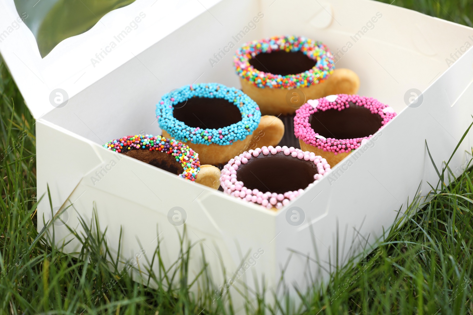 Photo of Box of delicious edible biscuit coffee cups decorated with sprinkles on green grass outdoors, closeup