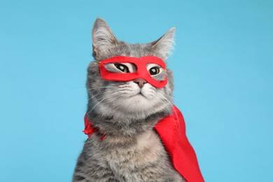Photo of Adorable cat in red superhero cape and mask on light blue background