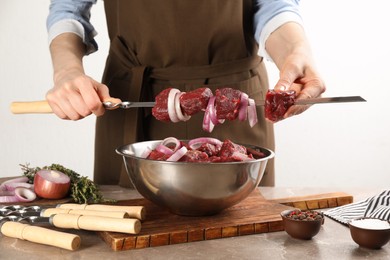 Photo of Woman stringing marinated meat and onion on skewer at table, closeup