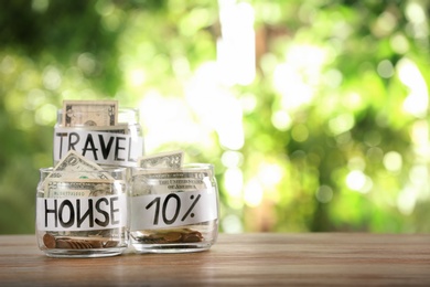 Glass jars with money for different needs on table against blurred background, space for text