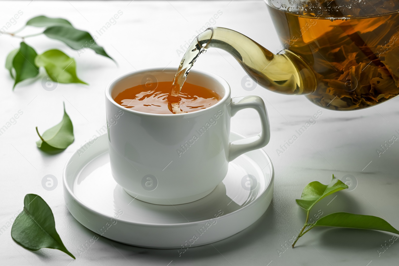 Photo of Pouring green tea into cup with saucer on white marble table, closeup
