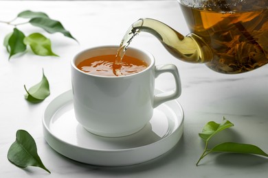 Pouring green tea into cup with saucer on white marble table, closeup
