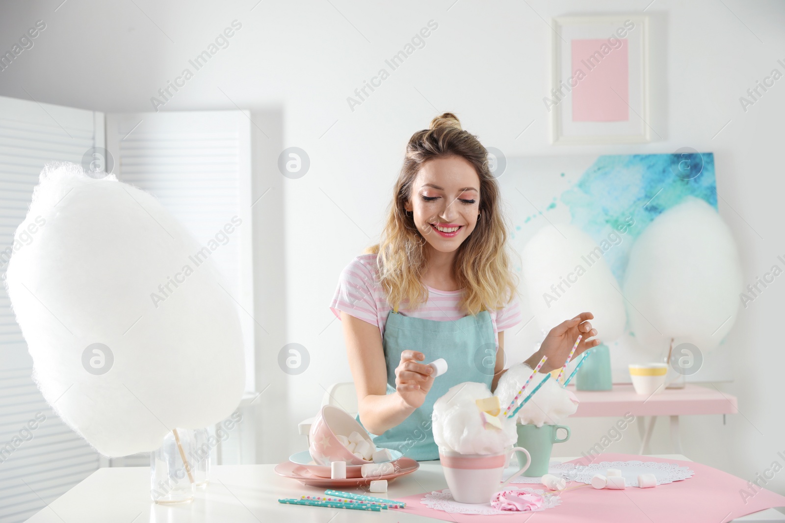 Photo of Young woman decorating cotton candy dessert with marshmallow at table in room