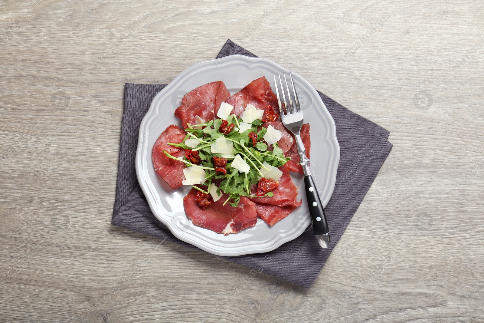 Photo of Plate of tasty bresaola salad with sun-dried tomatoes, parmesan cheese and fork on wooden table, top view