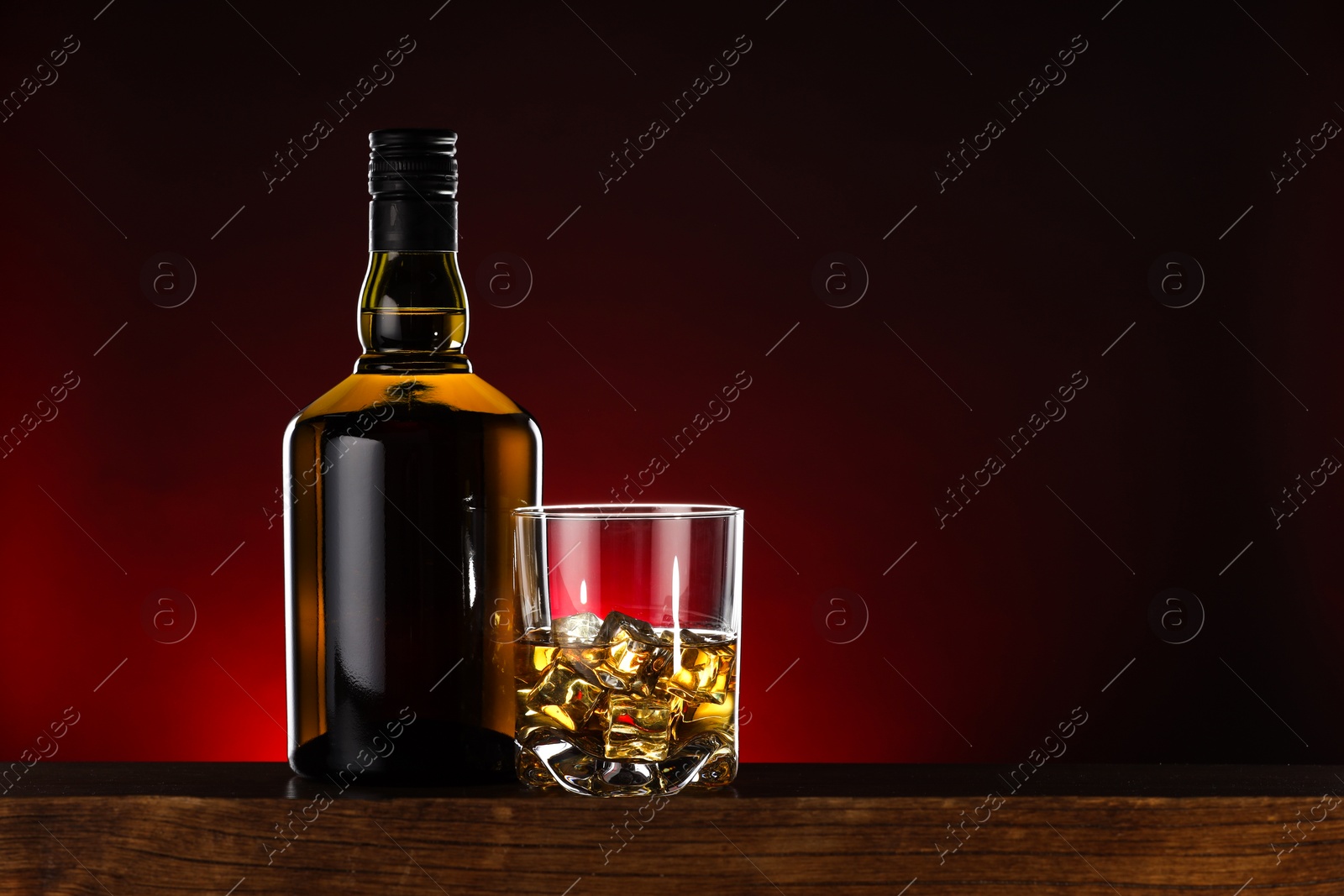 Photo of Whiskey with ice cubes in glass and bottle on wooden table, space for text