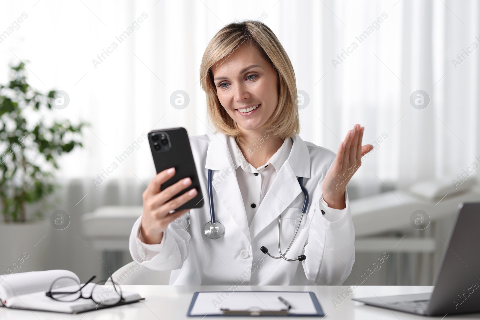 Photo of Smiling doctor with smartphone having online consultation at table in office