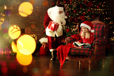 Photo of Santa Claus sneaking in with gift while little girl reading book near Christmas tree indoors