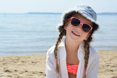 Little girl wearing sunglasses and hat at beach on sunny day. Space for text