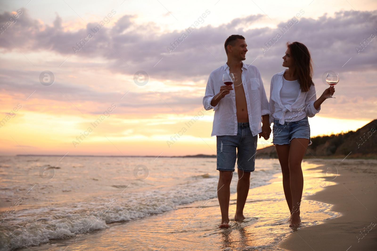 Photo of Lovely couple with glasses of wine walking on beach at sunset. Space for text