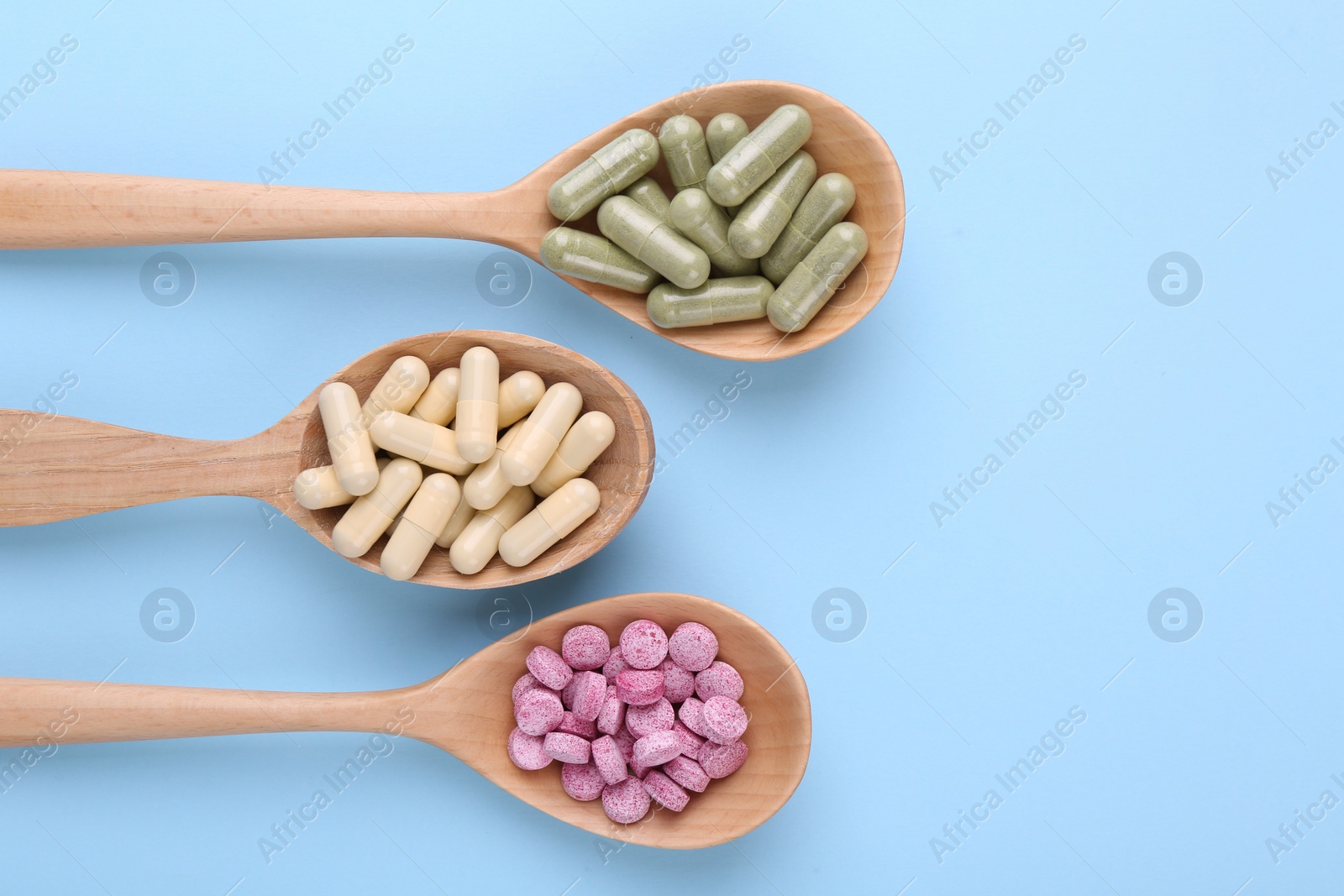 Photo of Different vitamin pills in wooden spoons on light blue background, flat lay. Space for text