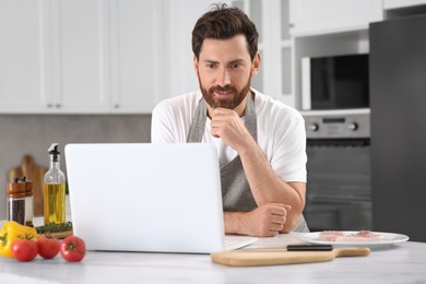 Photo of Man making dinner while watching online cooking course via laptop in kitchen