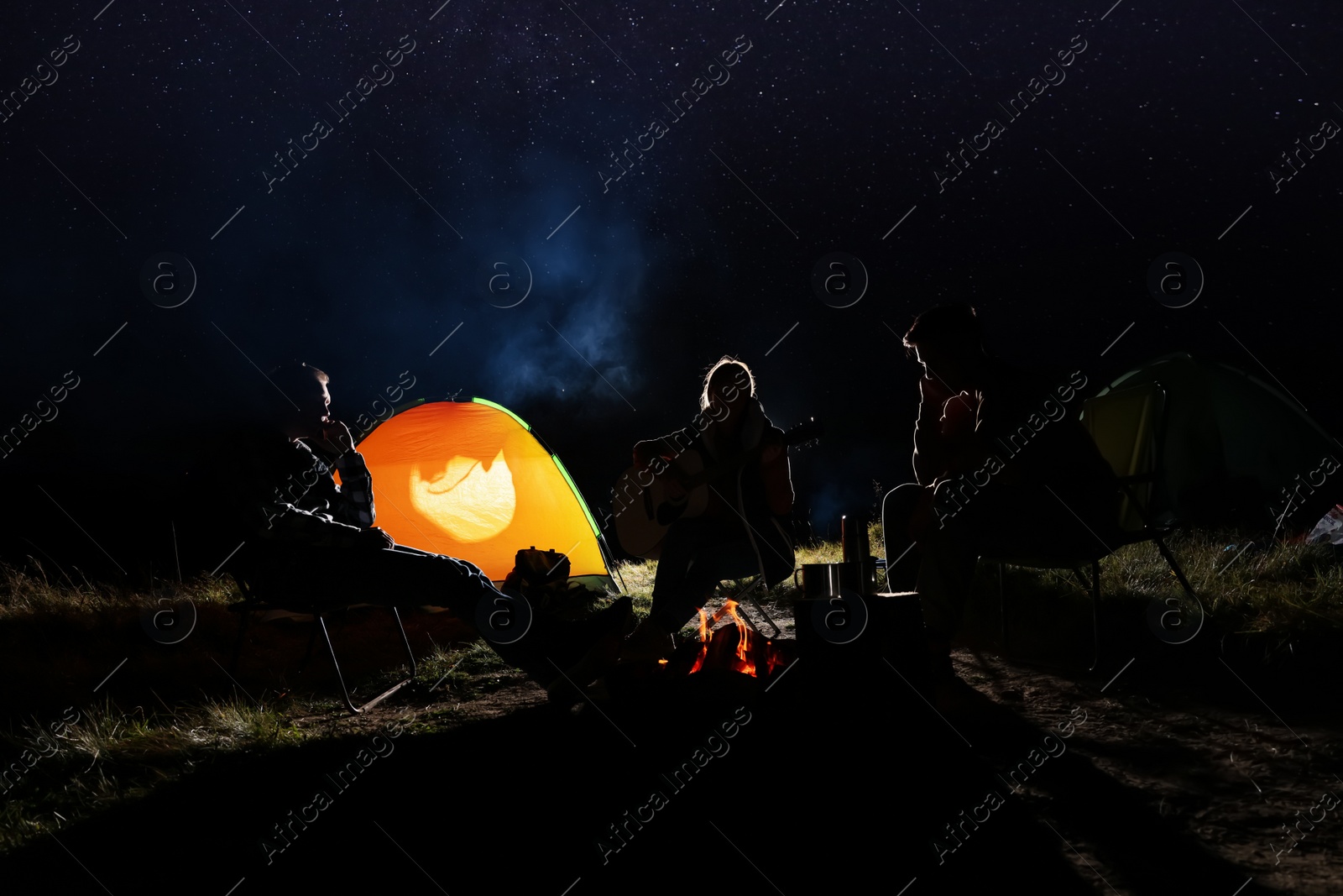 Photo of Group of friends with guitar near bonfire and camping tent outdoors at night