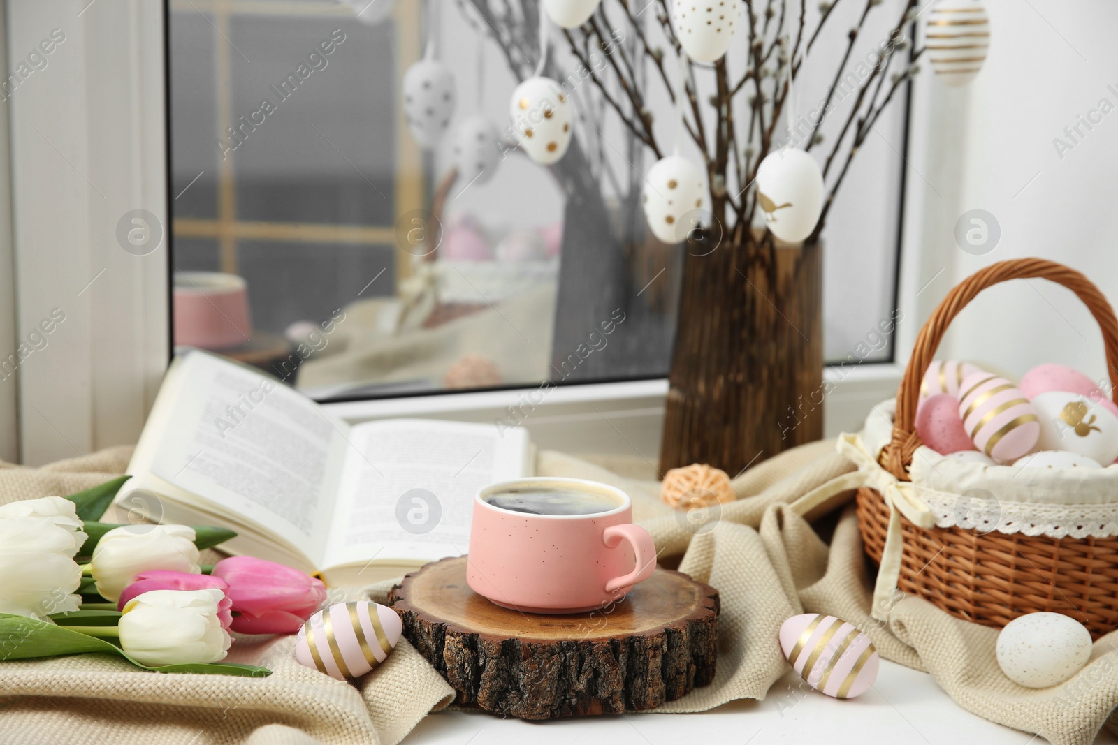 Photo of Easter decorations. Cup of coffee, painted eggs, tulips and book on windowsill