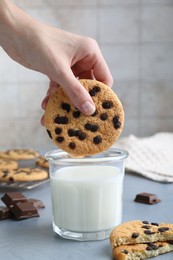 Photo of Woman dipping delicious chocolate chip cookie into glass of milk at grey table, closeup