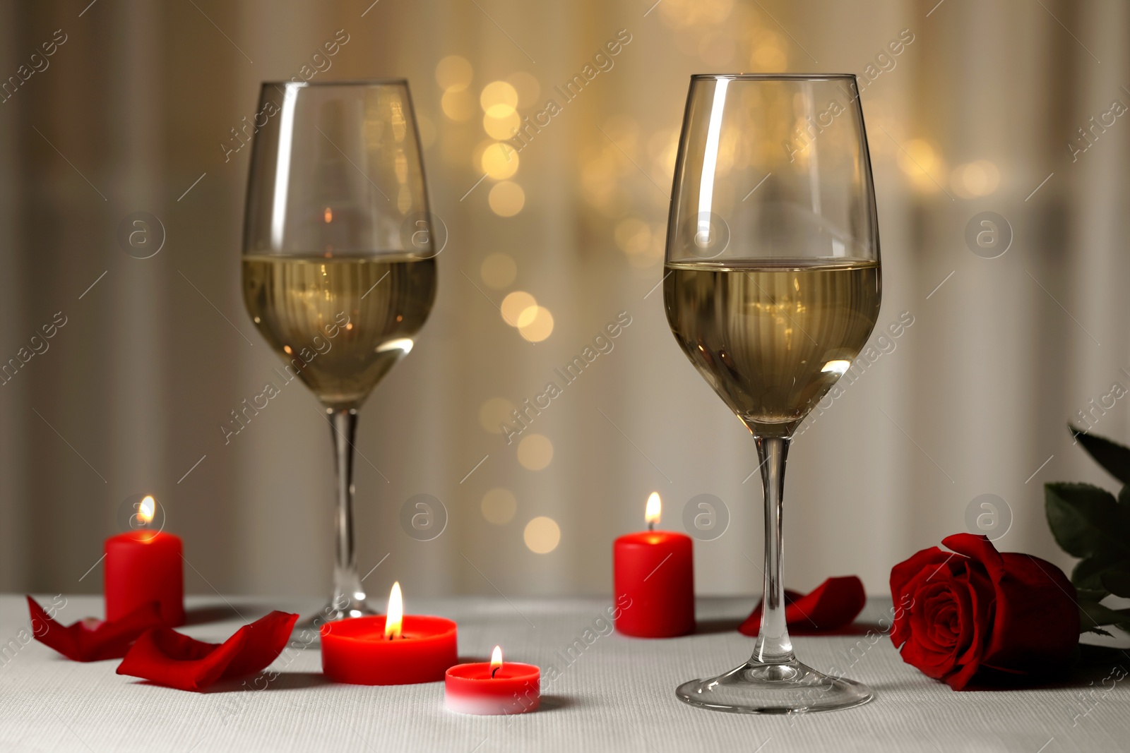 Photo of Glasses of white wine, rose flower and burning candles on grey table against blurred lights. Romantic atmosphere