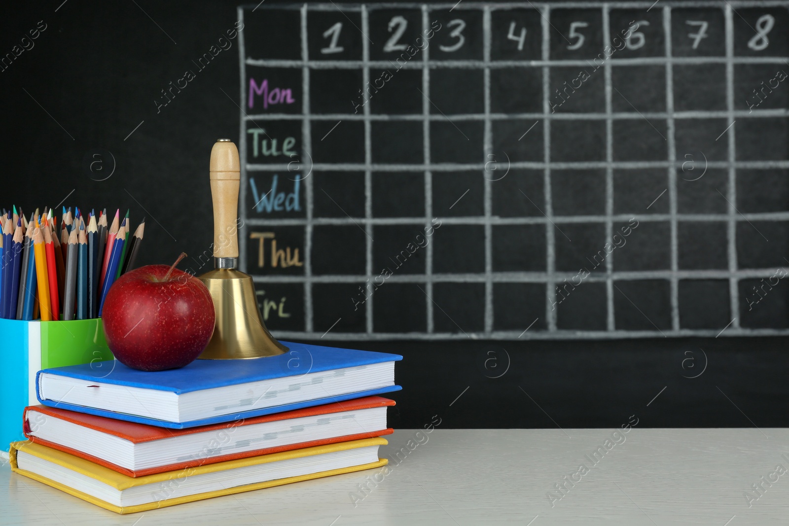 Photo of Apple, bell and different stationery on white wooden table near blackboard with chalked school timetable