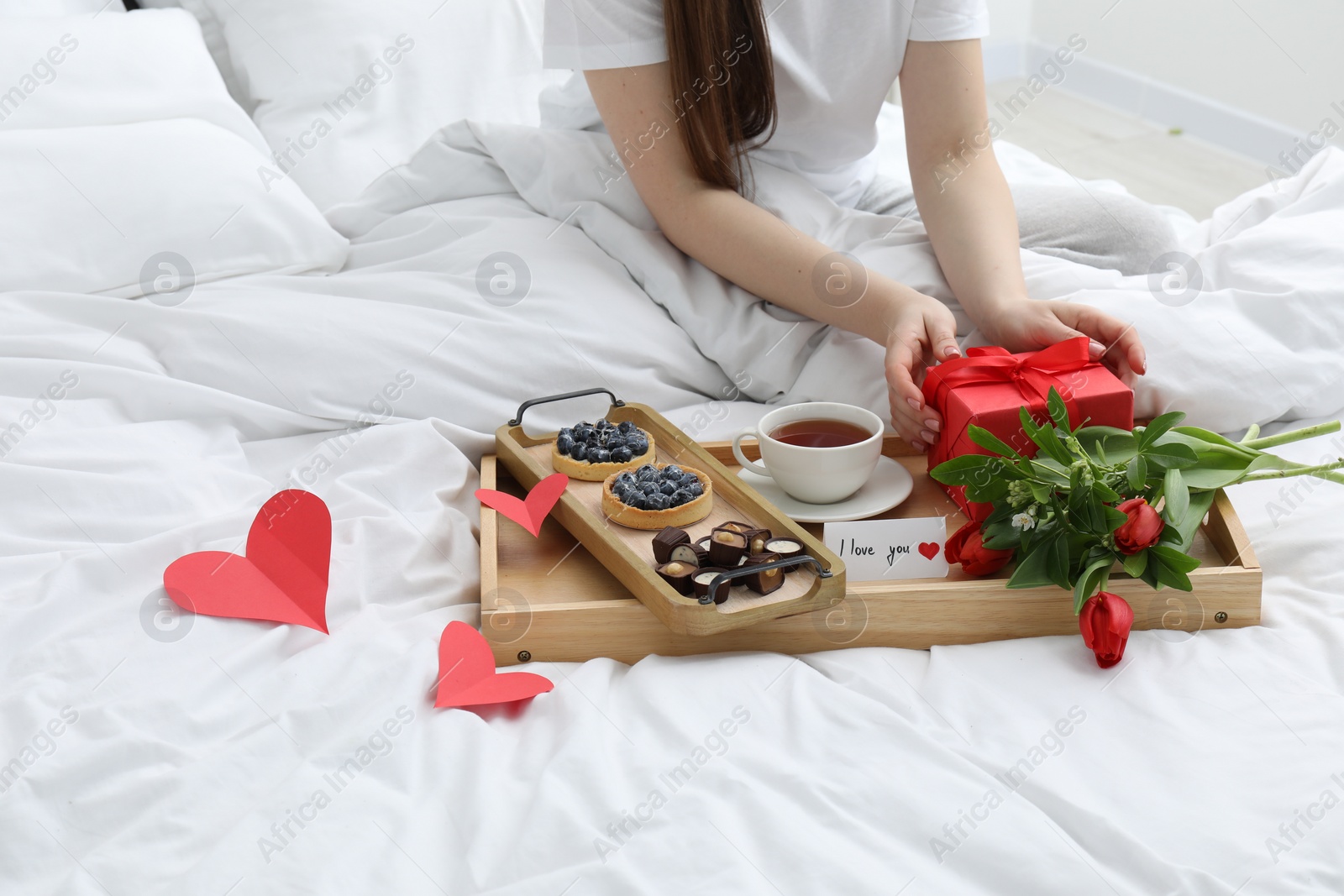 Photo of Tasty breakfast served in bed. Woman with gift box, tea, desserts, flowers and I Love You card at home, closeup